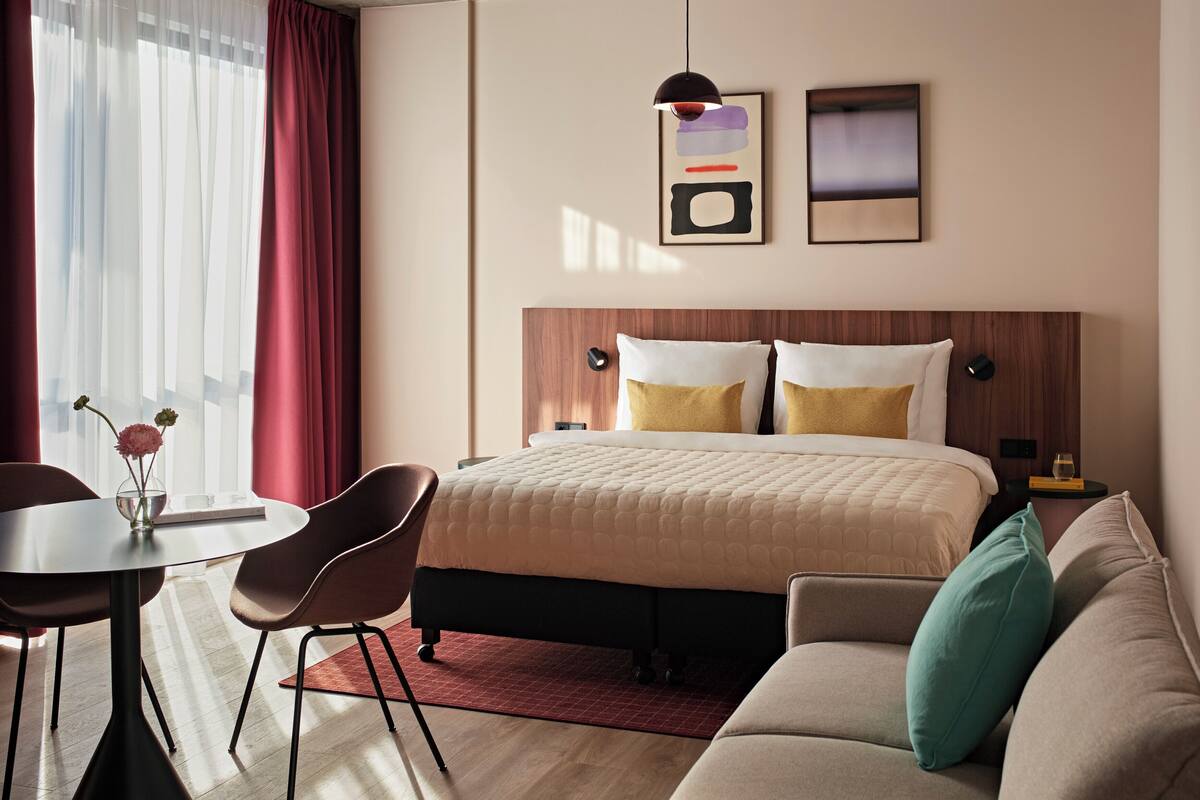 A modern hotel room with a bed, a sofa, a dining table with chairs, a vase with flowers, and artwork on the wall.