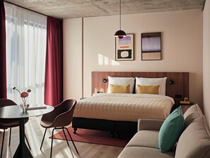 Ett modernt hotellrum med en säng, en soffa, ett matbord med stolar, en vas med blommor och konstverk på väggen.