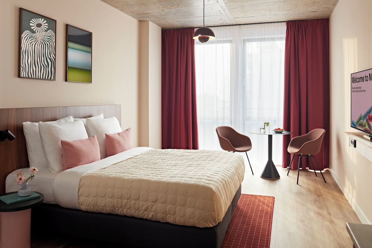 A hotel room with a bed, two chairs, a small table, a TV, and artwork on the wall.