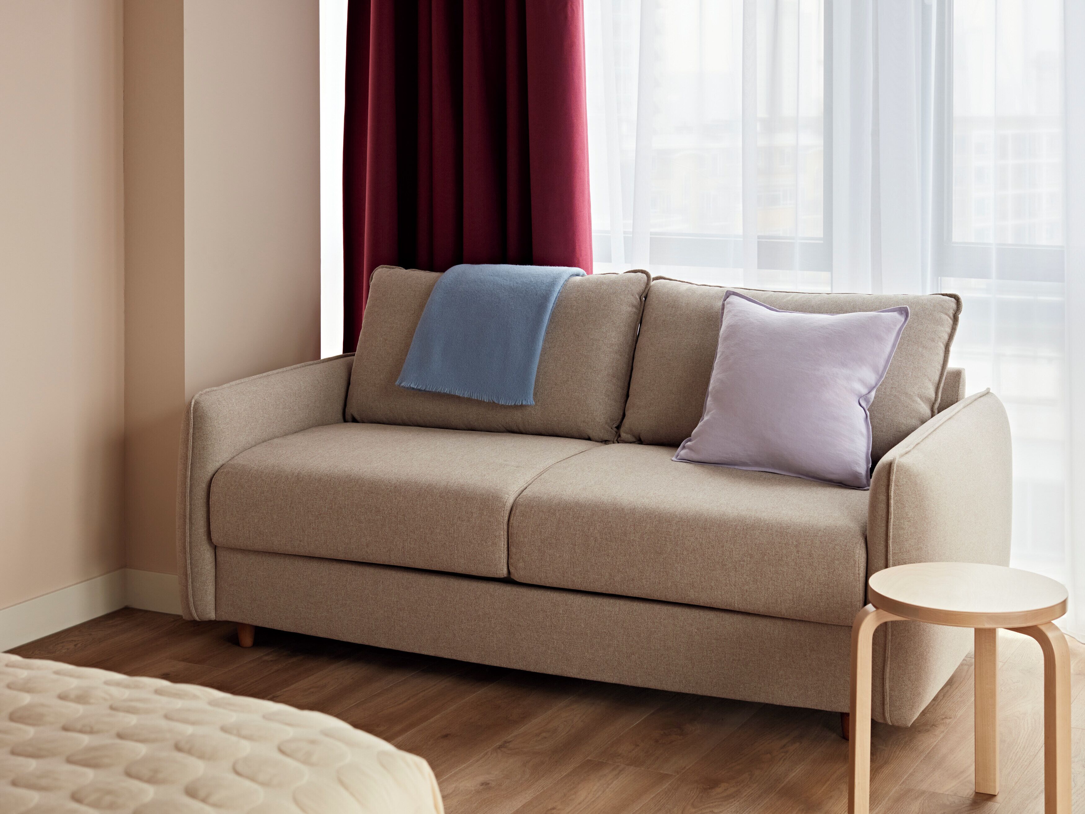 A beige sofa with two pillows, a small wooden stool, and a window with sheer curtains.