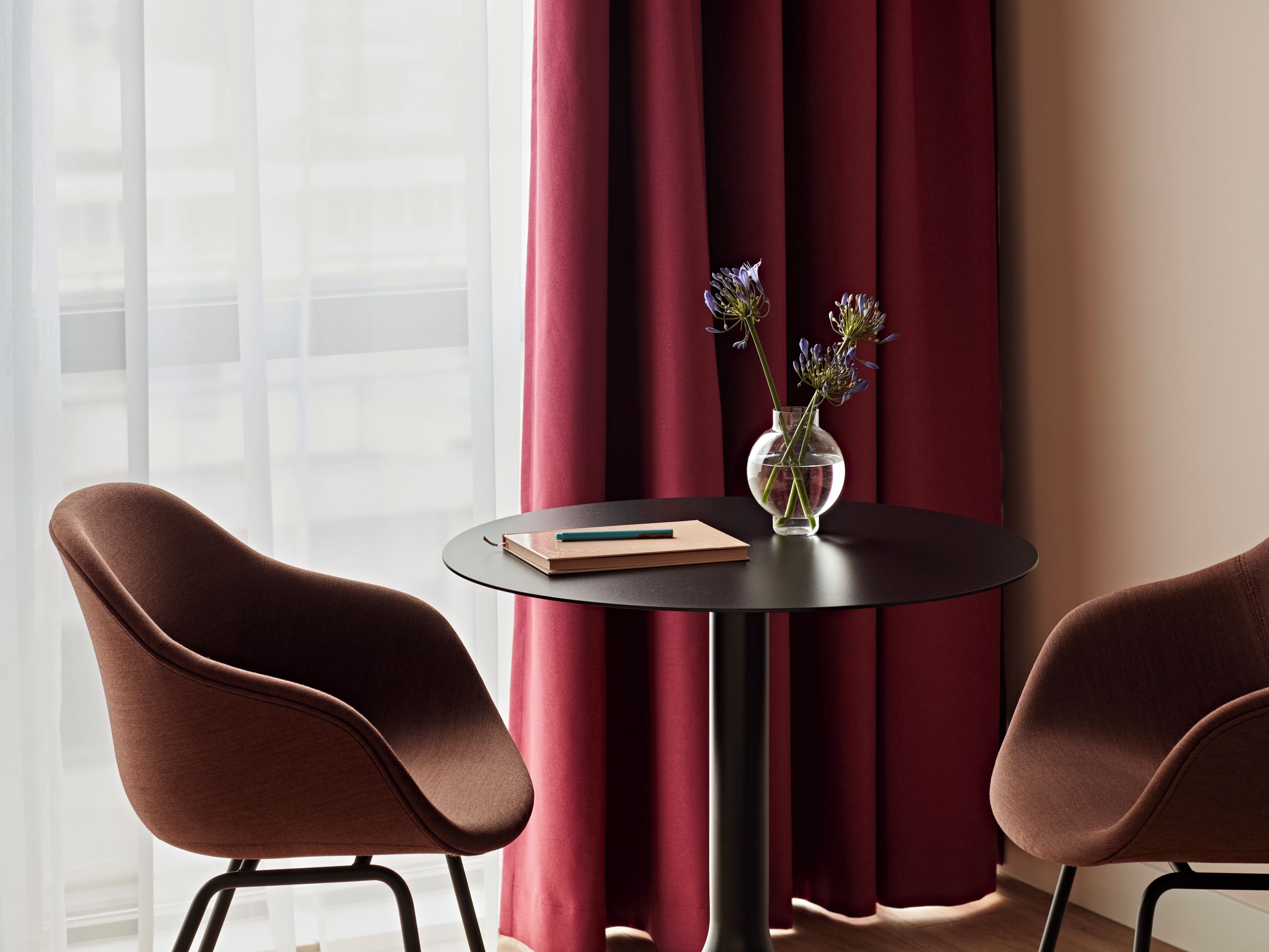Ein Zimmer mit zwei braunen Sesseln, einem kleinen runden Tisch mit einem Buch und einer Vase mit Blumen, sowie einem Fenster mit durchsichtigen Vorhängen.
