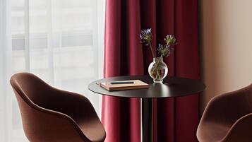 Ein Zimmer mit zwei braunen Sesseln, einem kleinen runden Tisch mit einem Buch und einer Vase mit Blumen, sowie einem Fenster mit durchsichtigen Vorhängen.