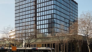 Ein modernes Hochhaus mit einem Raster aus Fenstern und einer Straßenbahnlinie vor der Fassade. Ein Schild mit der Aufschrift 'Fair Play' ist ebenfalls zu sehen.