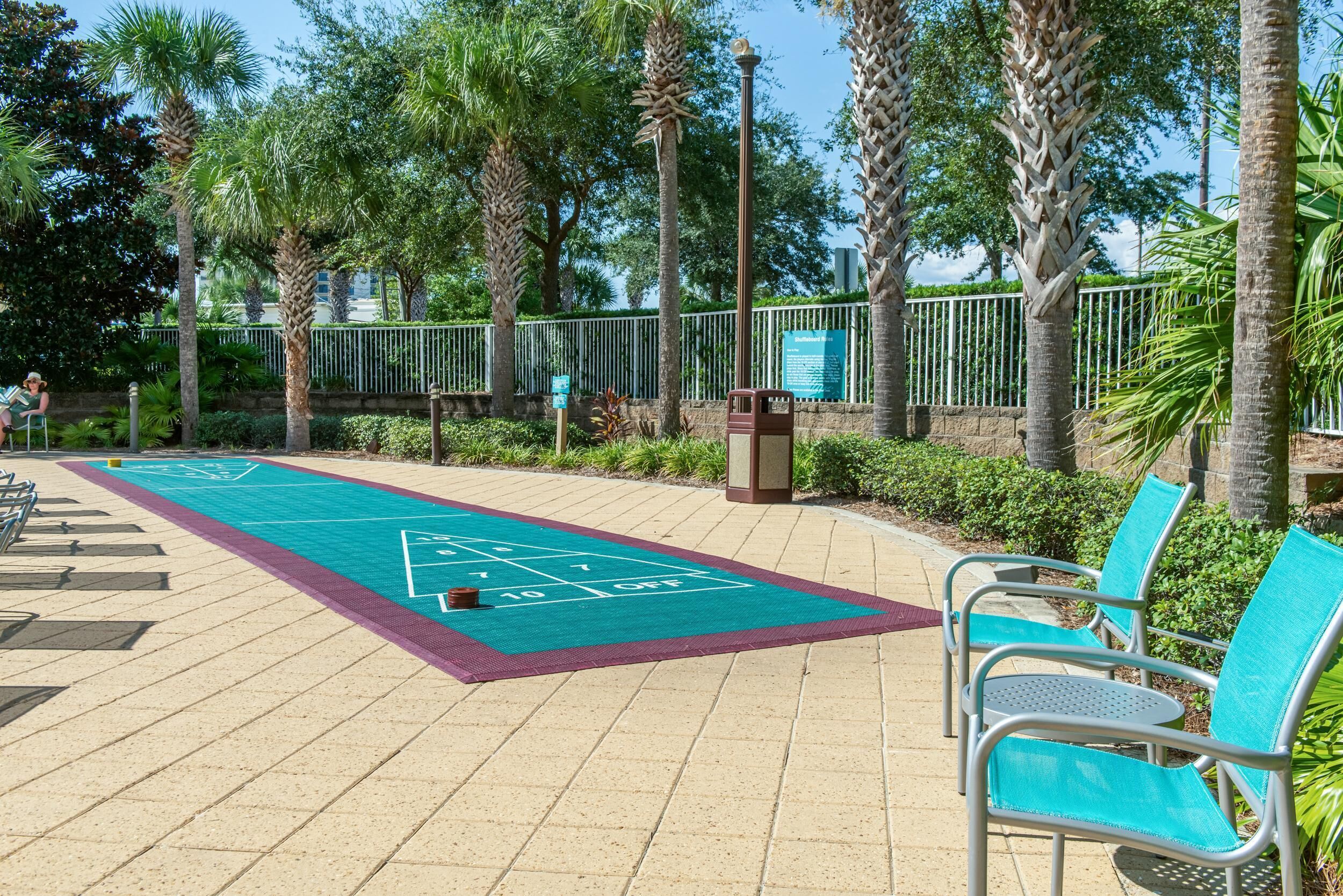 A sunny outdoor area with a shuffleboard court, metal chairs, and palm trees.