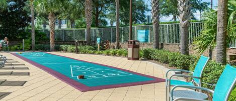 A sunny outdoor area with a shuffleboard court, metal chairs, and palm trees.