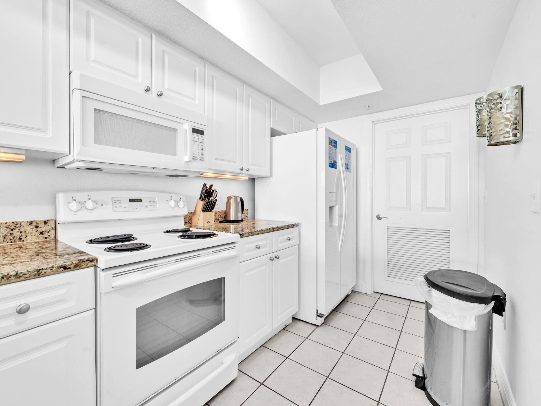 Una cocina moderna con gabinetes blancos, microondas, estufa, refrigerador y un bote de basura.