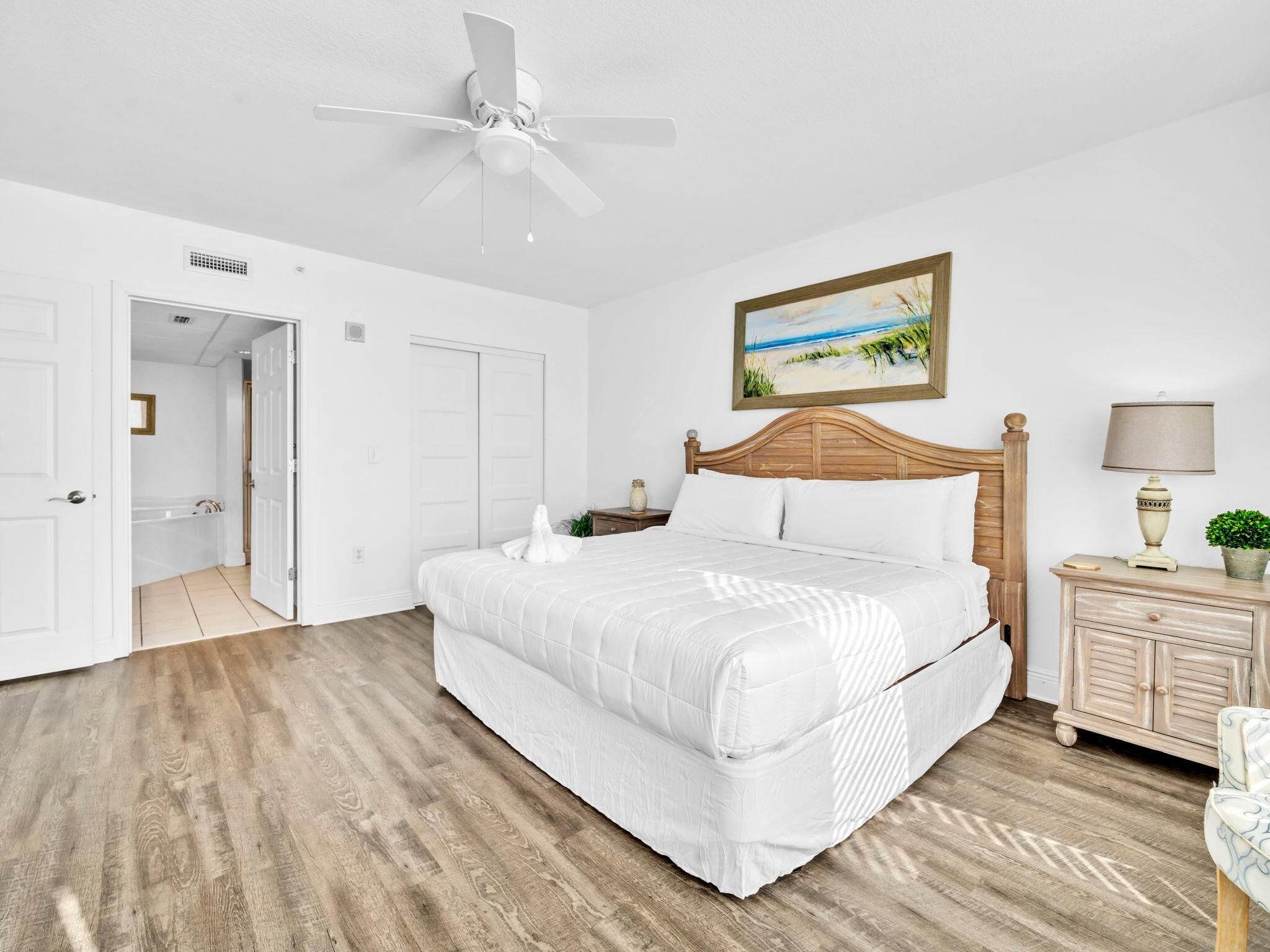 A bedroom with a wooden bed, white bedding, a ceiling fan, a nightstand with a lamp, and a painting on the wall.
