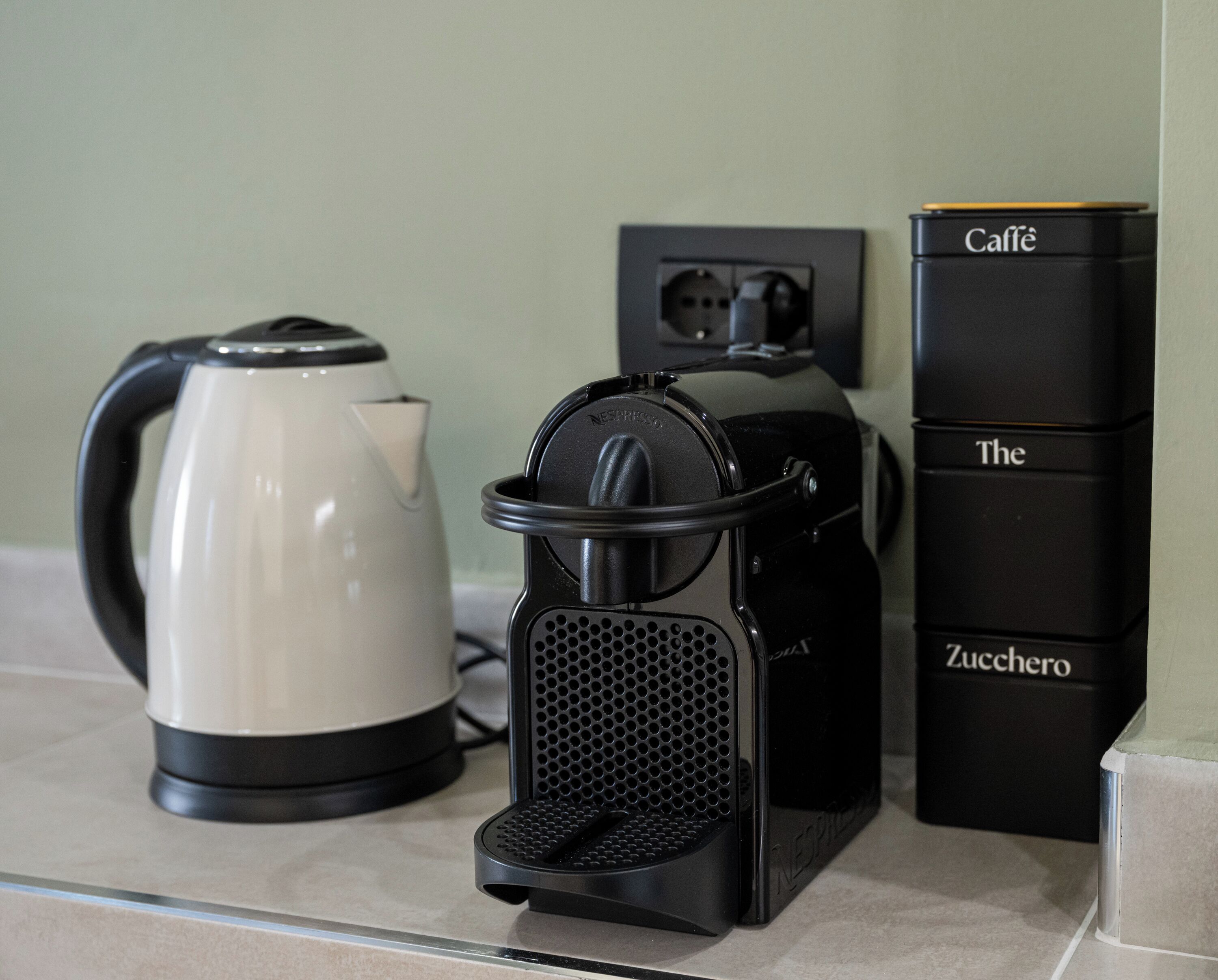 A kitchen counter with a coffee maker, a kettle, and a stack of black boxes labeled "Caffè The Zucchero."