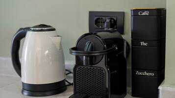 A kitchen counter with a coffee maker, a kettle, and a stack of black boxes labeled "Caffè The Zucchero."