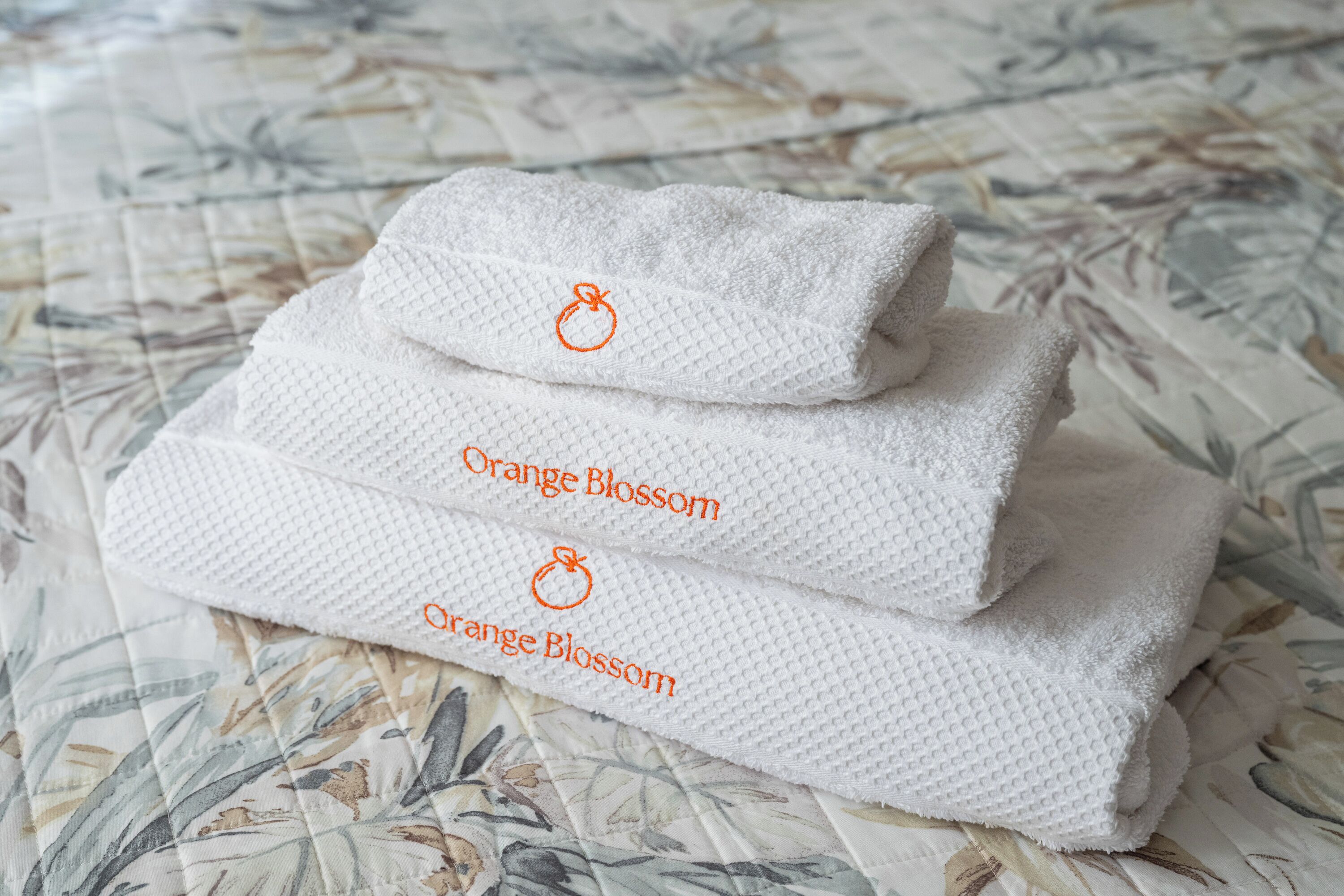 Three folded white towels with orange blossom embroidery on a floral-patterned bedspread.