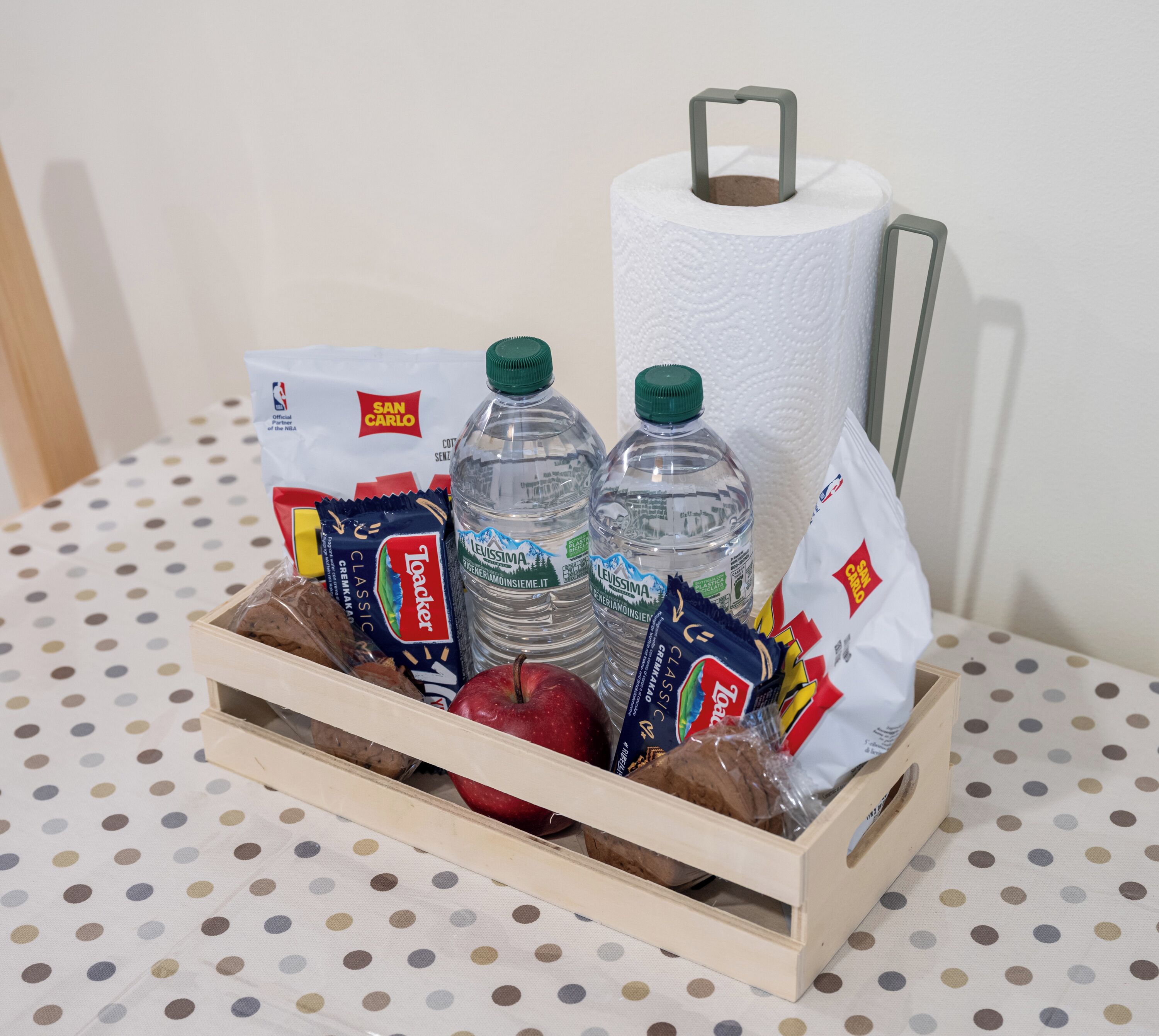 Een houten kist met een rol toiletpapier, twee waterflessen, een broodzak, een zak chips, een pak crackers en een appel.