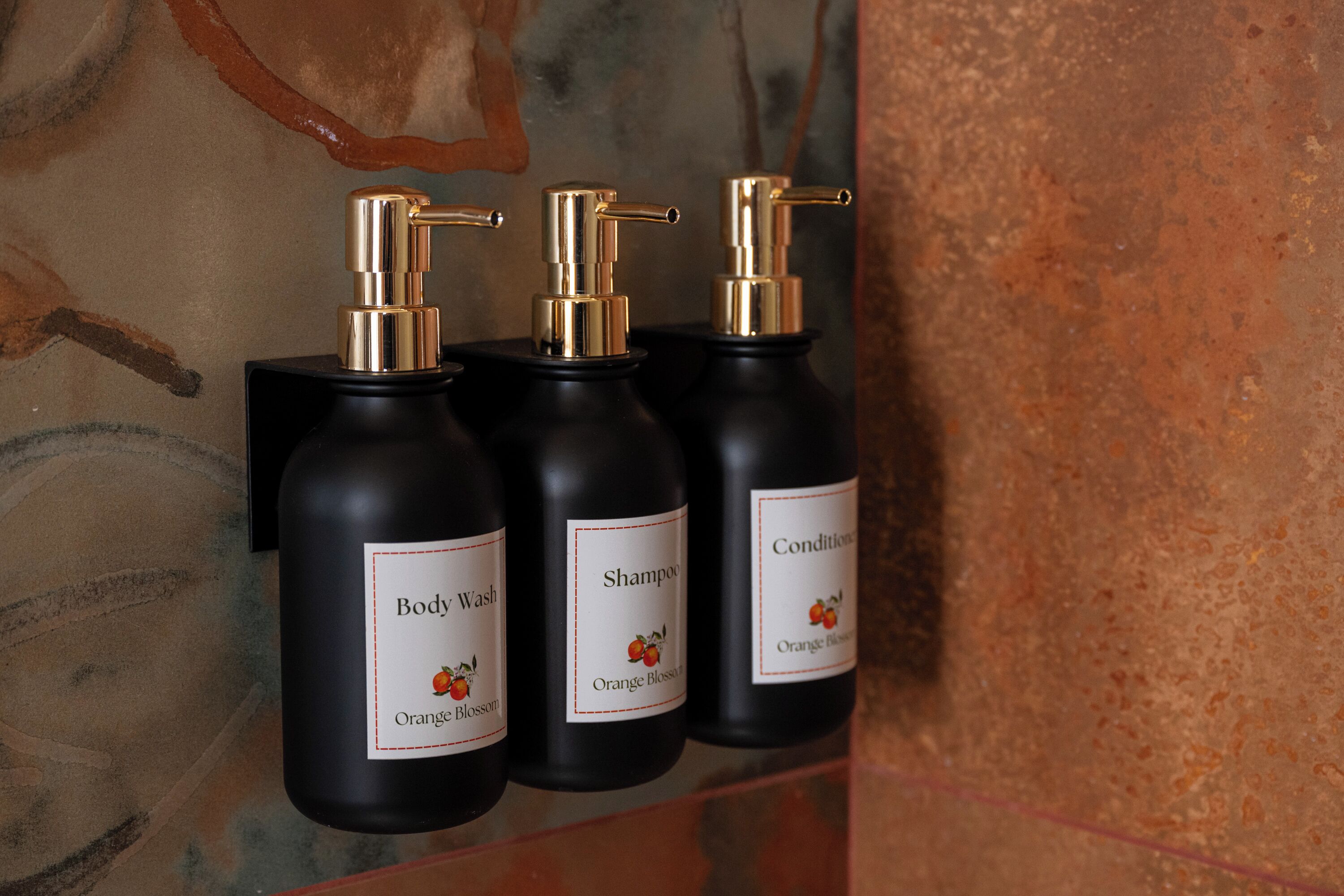 Drei schwarze Flaschen mit goldenen Dosierern, beschriftet mit 'Body Wash', 'Shampoo' und einer Flasche 'Conditioner', alle mit Orangeblüten-Duft.