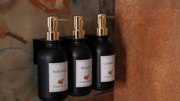 Drei schwarze Flaschen mit goldenen Dosierern, beschriftet mit 'Body Wash', 'Shampoo' und einer Flasche 'Conditioner', alle mit Orangeblüten-Duft.