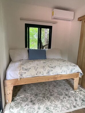 Uma cama de solteiro com colcha e almofadas estampadas com motivos florais, uma janela com vista para o verde e um aparelho de ar condicionado acima da janela.