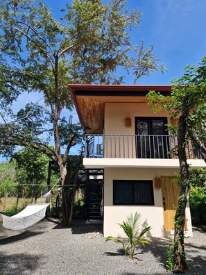 Uma casa moderna de dois andares com varanda, cercada por árvores e uma rede.