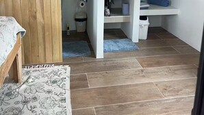 Une chambre à coucher avec une armoire en bois, un lit recouvert d’une couvre-lit à motifs floraux, et une salle de bain équipée d’un lavabo, de toilettes et d’un porte-serviettes.
