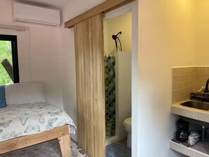 Quarto compacto com uma cama, guarda-roupa de madeira, chuveiro com porta decorada com padrão de folha e pequena cozinha equipada com pia e armários.