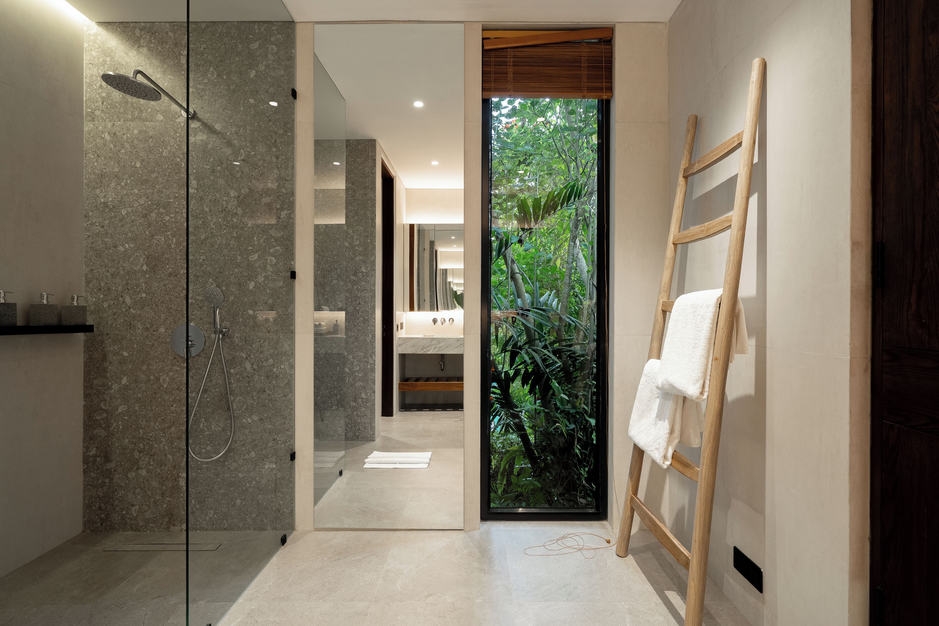 Uma casa de banho moderna com chuveiro de vidro, uma escada de madeira e vista para um jardim tropical através de uma porta de vidro.