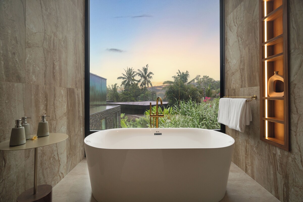 Un baño moderno con una bañera independiente, una mesita de noche con artículos de tocador y vistas a palmeras y al atardecer.