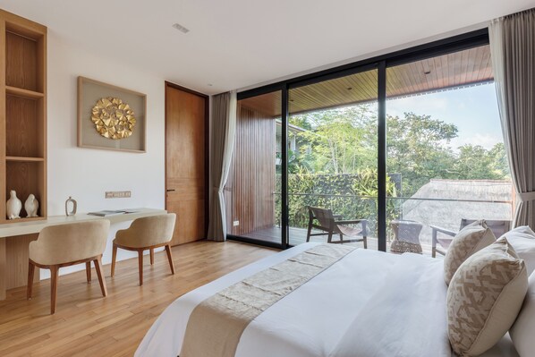 A modern bedroom with a large bed, a desk, two chairs, and a balcony with a view of greenery. - Seven Stones Villas (Ubud)
