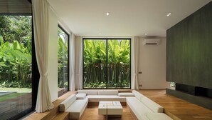 A modern living room with a large sliding glass door leading to a garden, a sectional sofa, a coffee table, and a dark accent wall. - Seven Stones Villas (Ubud)