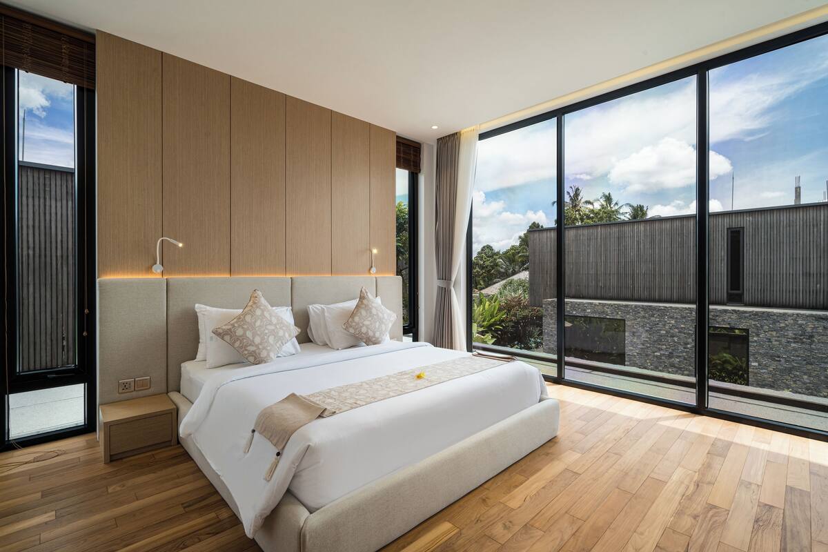 A modern bedroom with a large bed, wooden paneling, and a view of a garden and building through large windows.