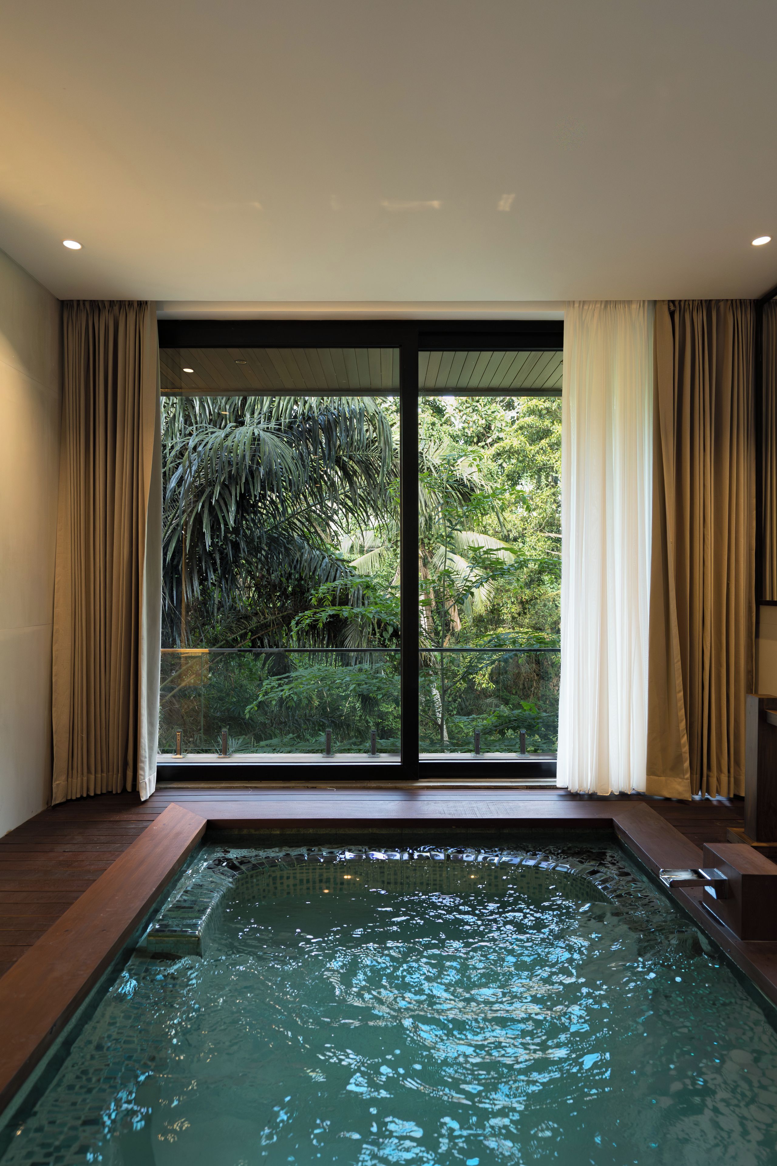 A modern indoor swimming pool with a glass barrier, surrounded by beige curtains and a view of lush greenery.