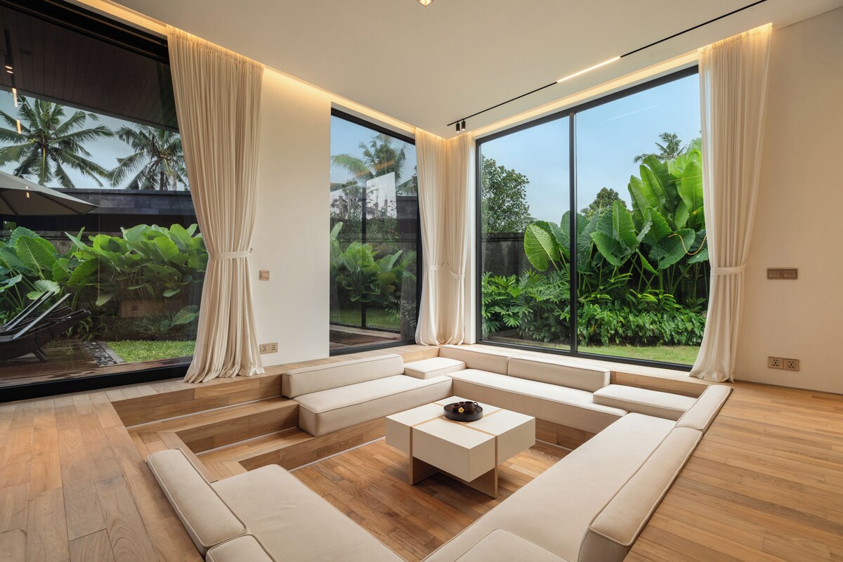 Una sala de estar moderna con un amplio sofá modular, una mesa de centro y vistas a exuberante vegetación a través de amplios ventanales.