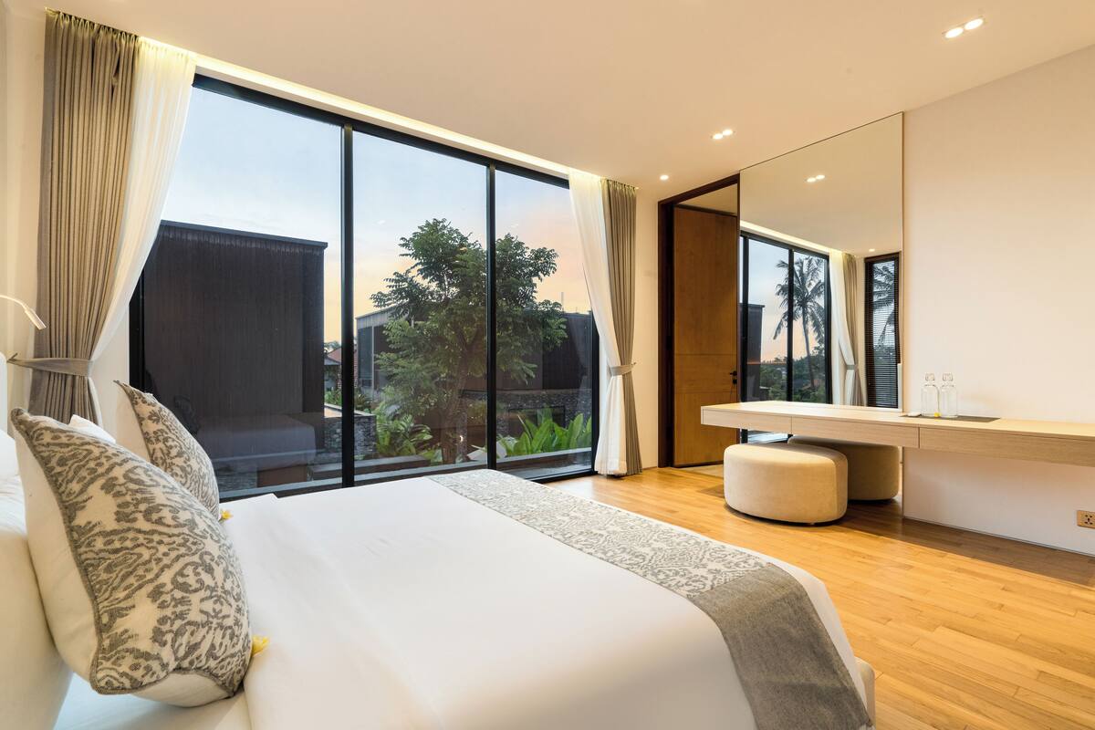 A modern bedroom with a large bed, a vanity with a mirror, and a view of the outdoors.
