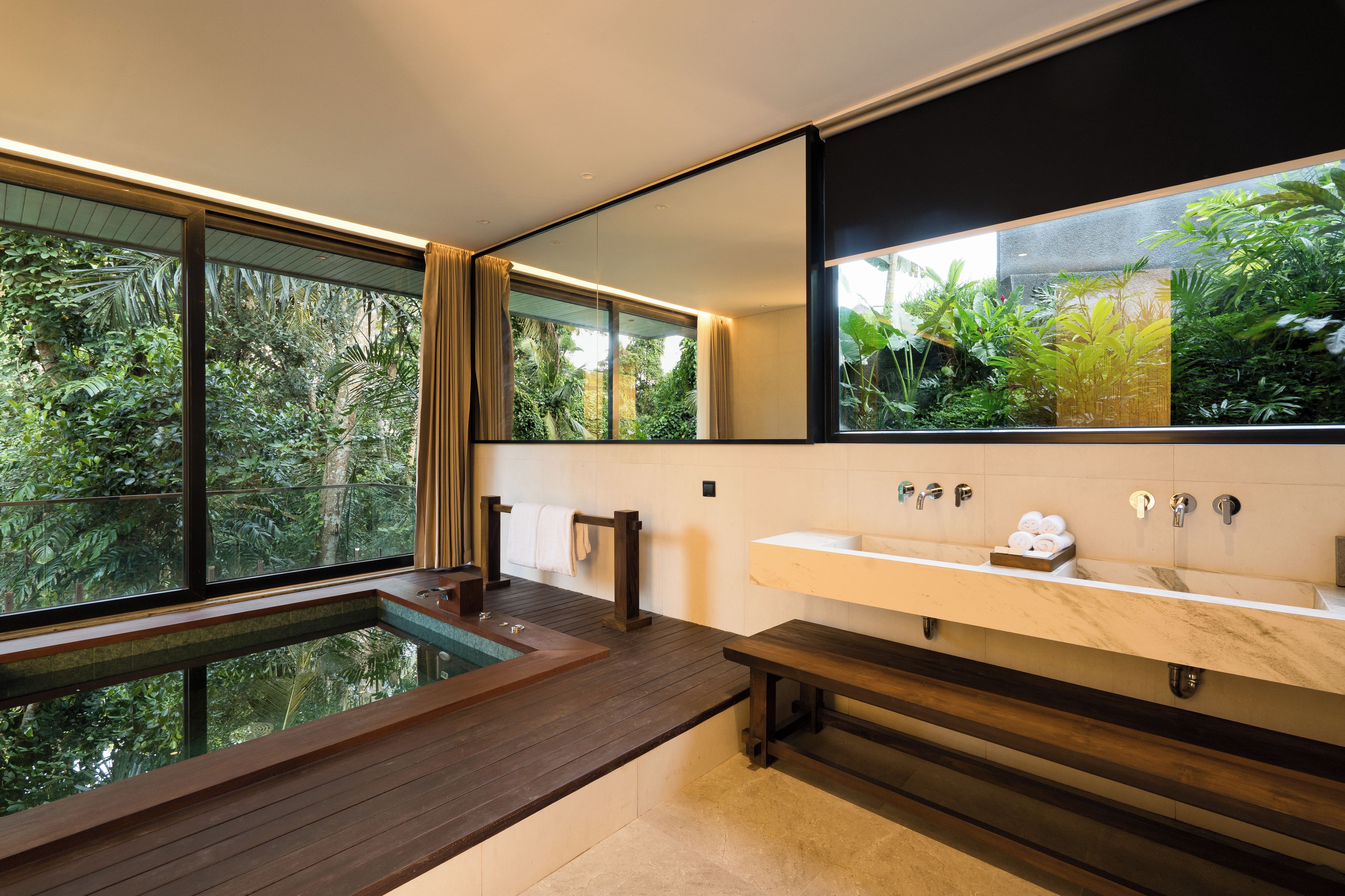 A modern bathroom with a wooden bench, a large mirror, and a view of lush greenery outside.
