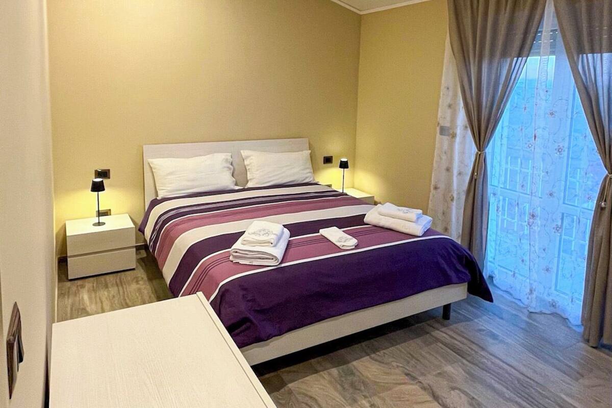 Un dormitorio con una cama, dos mesitas de noche, un tocador y un armario, junto a una ventana con cortinas.