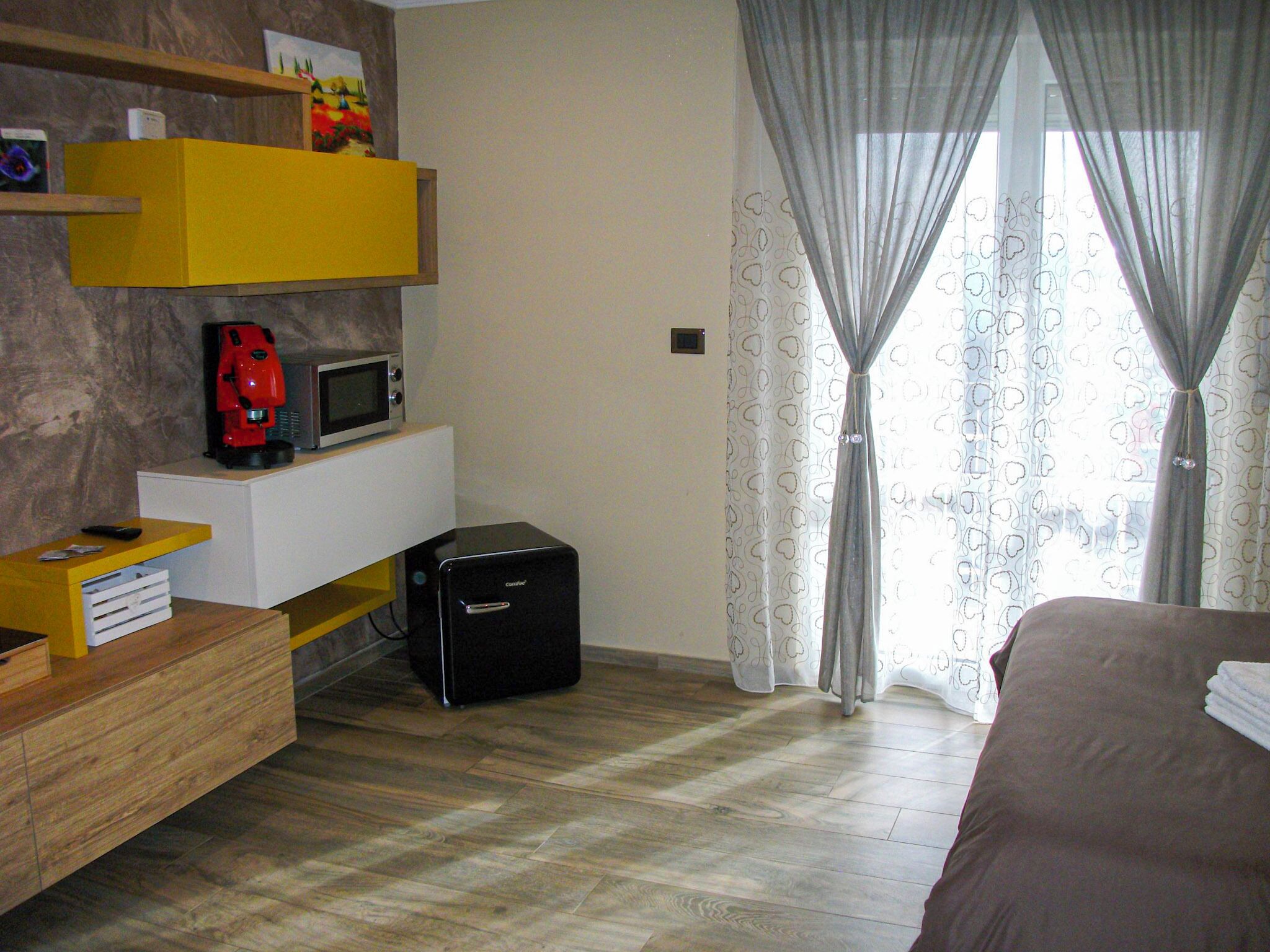 A bedroom with a bed, a desk with a coffee machine, a suitcase, and a window with curtains.