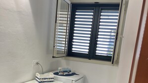 A compact kitchen with a white countertop, a small refrigerator, a window with blinds, and a colorful storage unit under the counter. - LUISA ROOM (Sampieri)