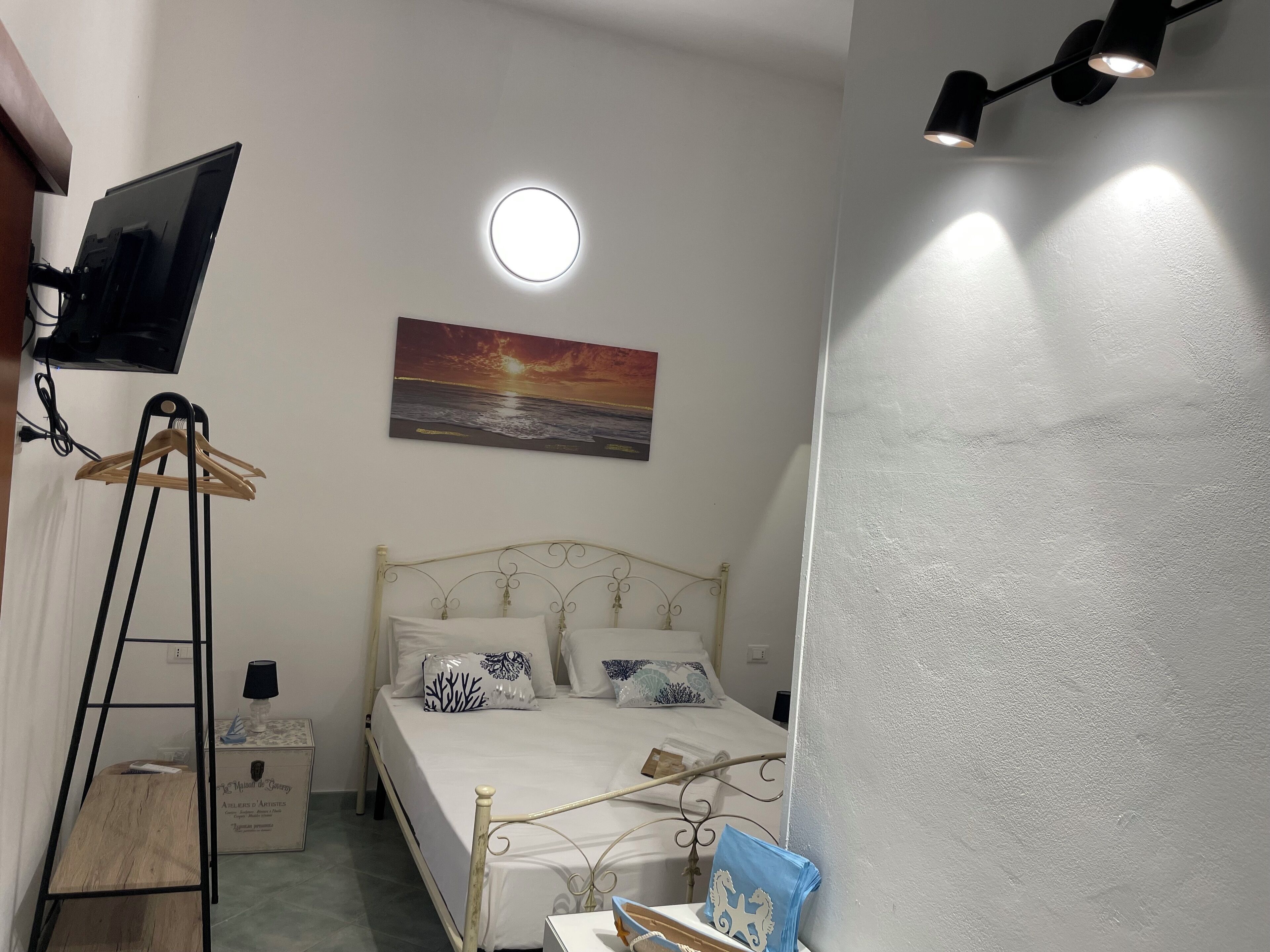 Una camera da letto con un letto, una TV a parete, un quadro raffigurante un tramonto e un comodino con una lampada.