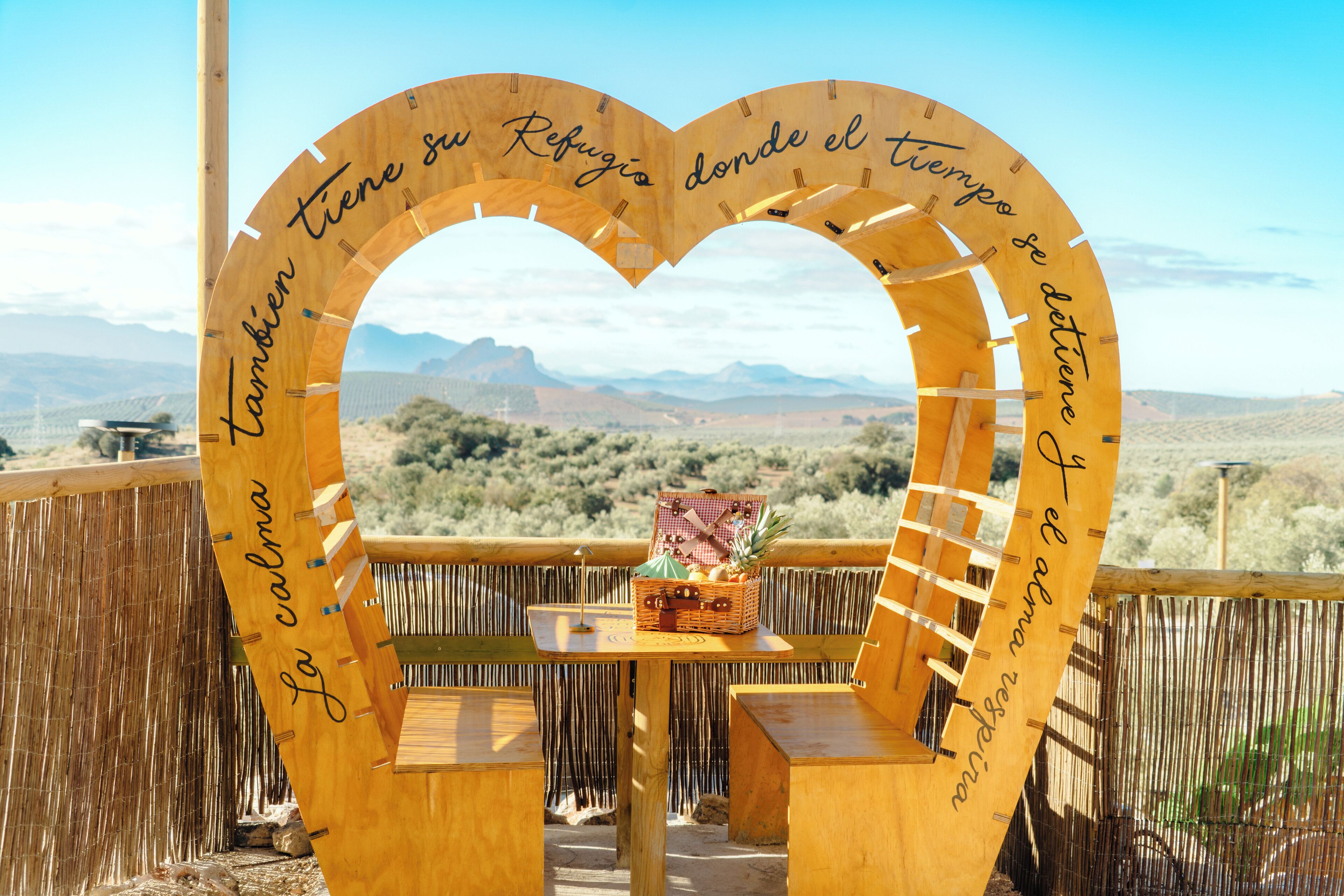 Una estructura de madera con forma de corazón, una mesa con una cesta y vistas a montañas.