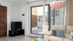 A modern living room with a television on a black stand, a beige sofa with cushions, a glass coffee table with a plant, and a large window with curtains. - Sliema Seaside Penthouse with Terrace (Sliema)