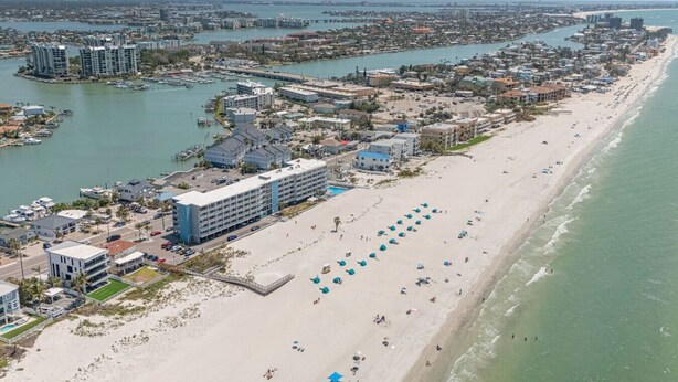 Ein Luftbild einer Küstenstadt mit einem langen Sandstrand, einem Jachthafen und mehreren Gebäuden, darunter ein Hotel mit Pool und Strandstühlen.