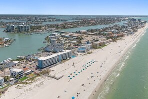 Vue aérienne d’une ville côtière avec une longue plage de sable, un port de plaisance et plusieurs bâtiments, dont un hôtel doté d’une piscine et de parasols de plage.