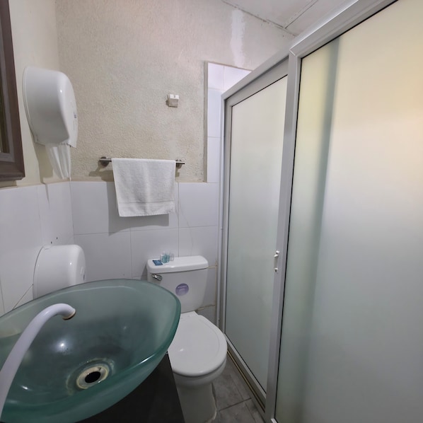 Un baño con lavamanos de vidrio, inodoro, toalla colgada en un perchero y puerta de ducha.