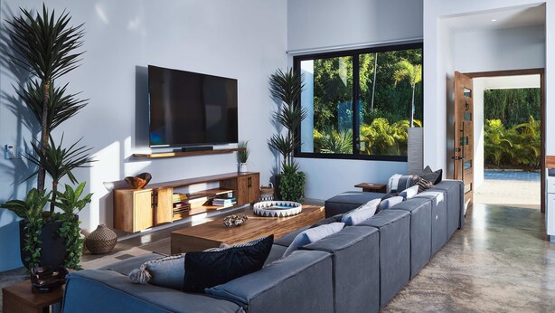 Una sala de estar moderna con un televisor grande, un sofá y una mesa de centro. Hay plantas y un mueble de madera.