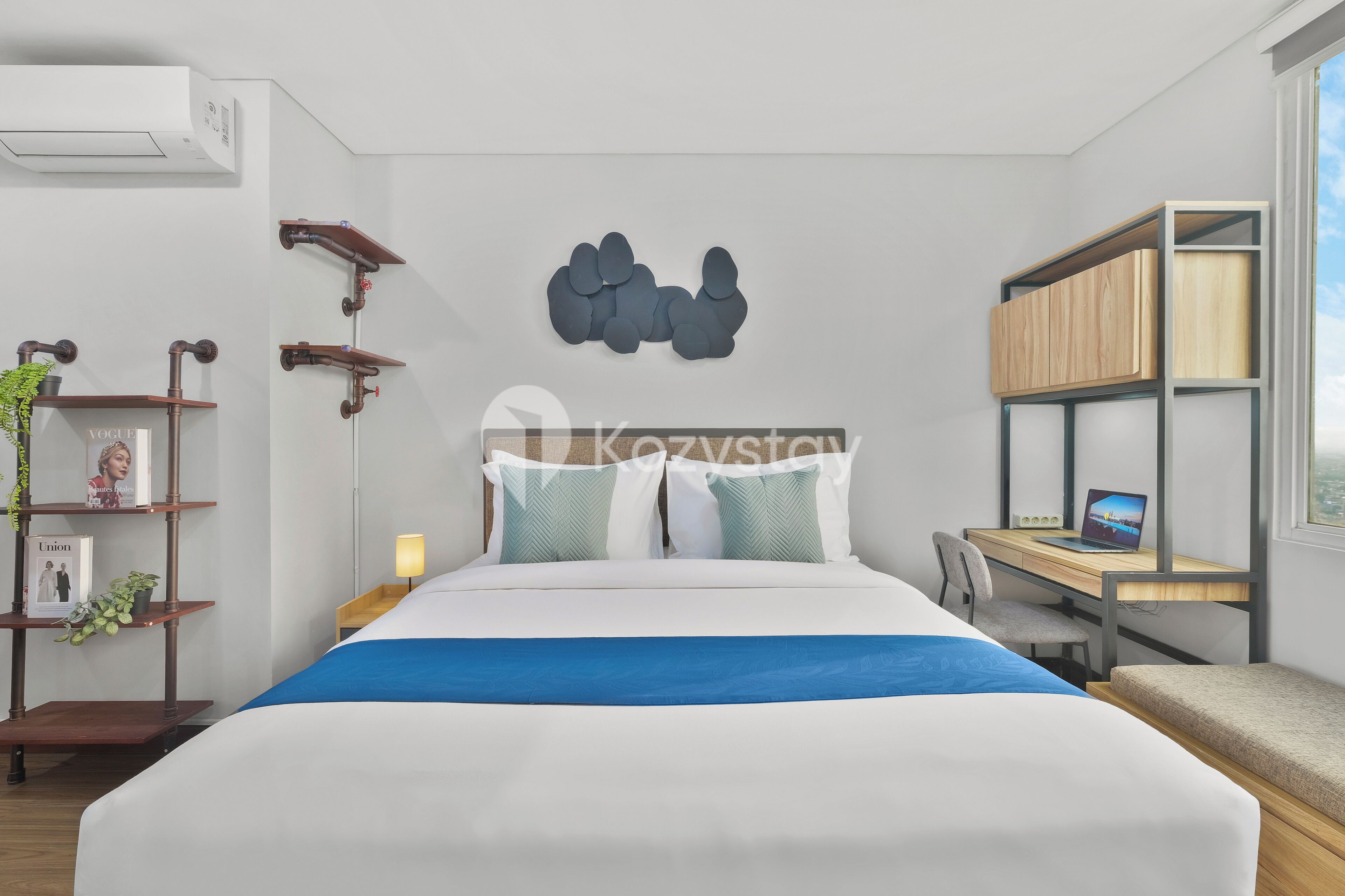 A modern hotel room with a large bed, a desk with a laptop, a bookshelf, and a window with a city view.