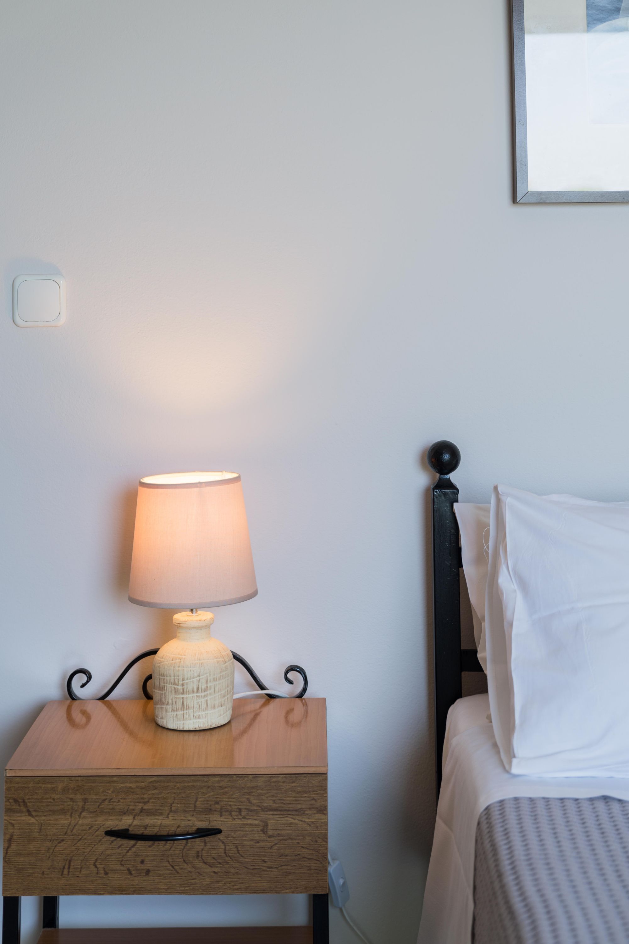 Ein Nachttisch mit Lampe, ein Bett mit weißer Bettwäsche und ein gerahmtes Bild an der Wand.