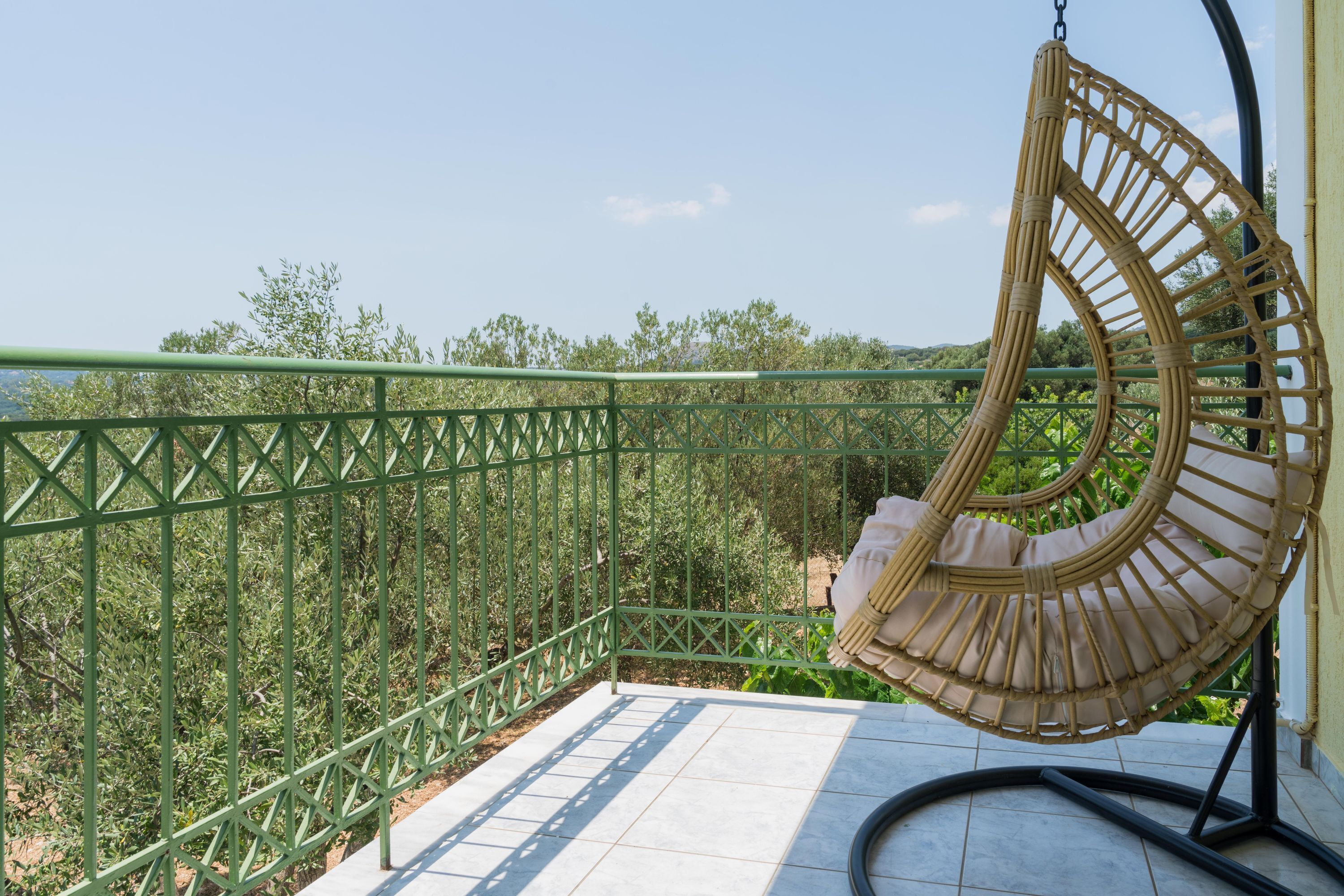 Una silla colgante de mimbre en un balcón con baranda de metal verde y vistas a la vegetación.