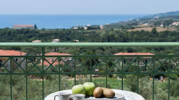 Varanda com mesa posta para dois, com prato de frutas e uma xícara, com vista para uma paisagem deslumbrante com casas e o mar.