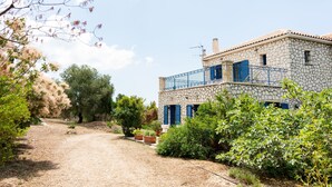 A stone house with blue shutters and a balcony, surrounded by greenery and a gravel path. - Stone House in Olive Grove (Kefalonia)