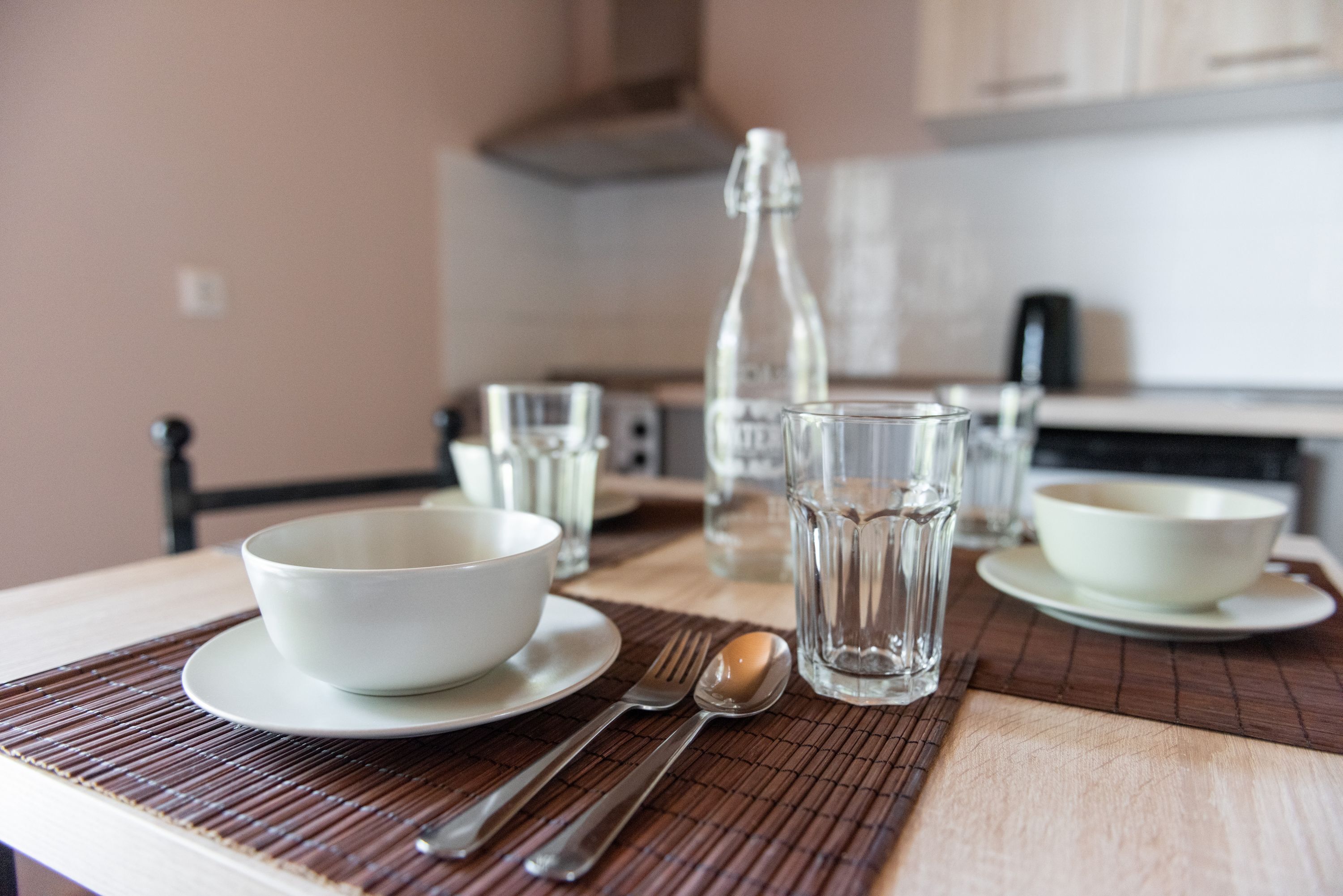 A dining table with a bowl, spoon, fork, and glasses on a bamboo placemat.