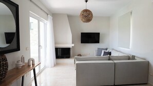 A modern living room with a sectional sofa, a flat-screen TV mounted on the wall, a fireplace, and a large window with curtains. - Vasilopoulos Residences - Villa Kalliste (Kefalonia)