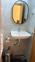 A bathroom with a white sink, a round mirror, and a stainless steel trash can.