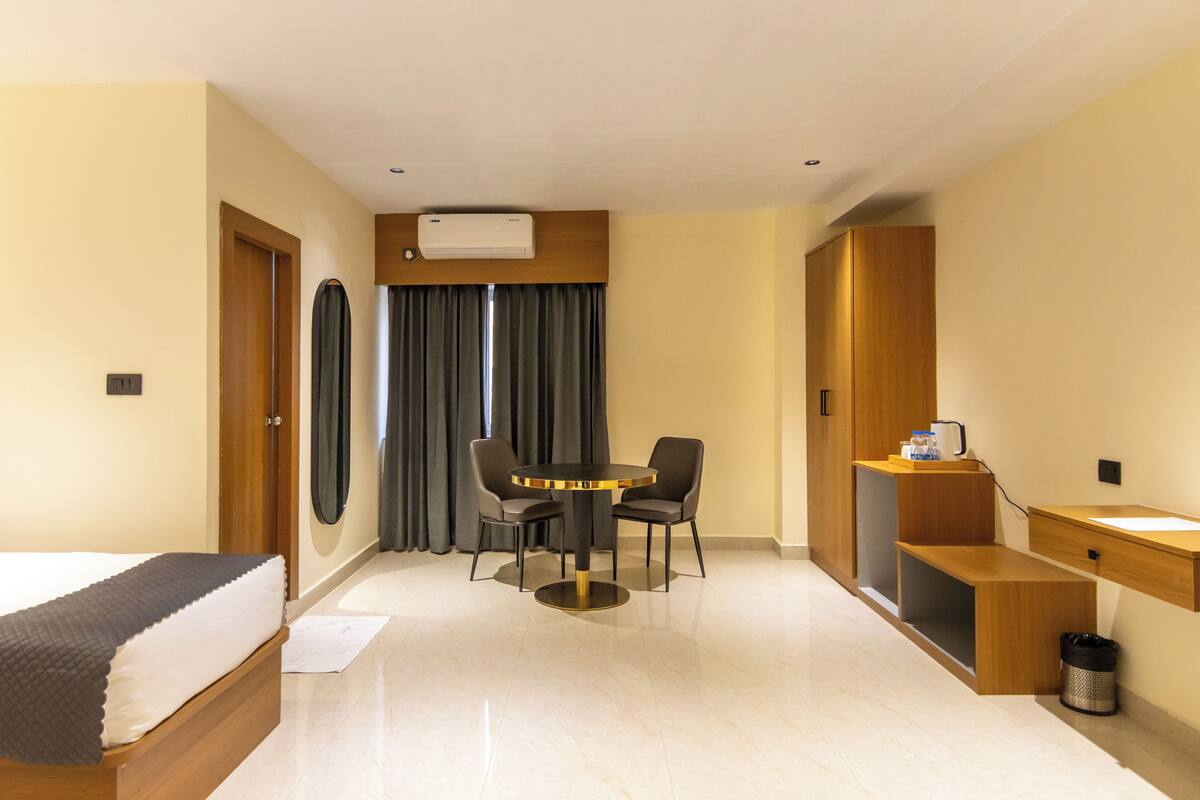 A hotel room with a bed, a round table with chairs, a desk with a chair, a small shelf, and a trash bin.