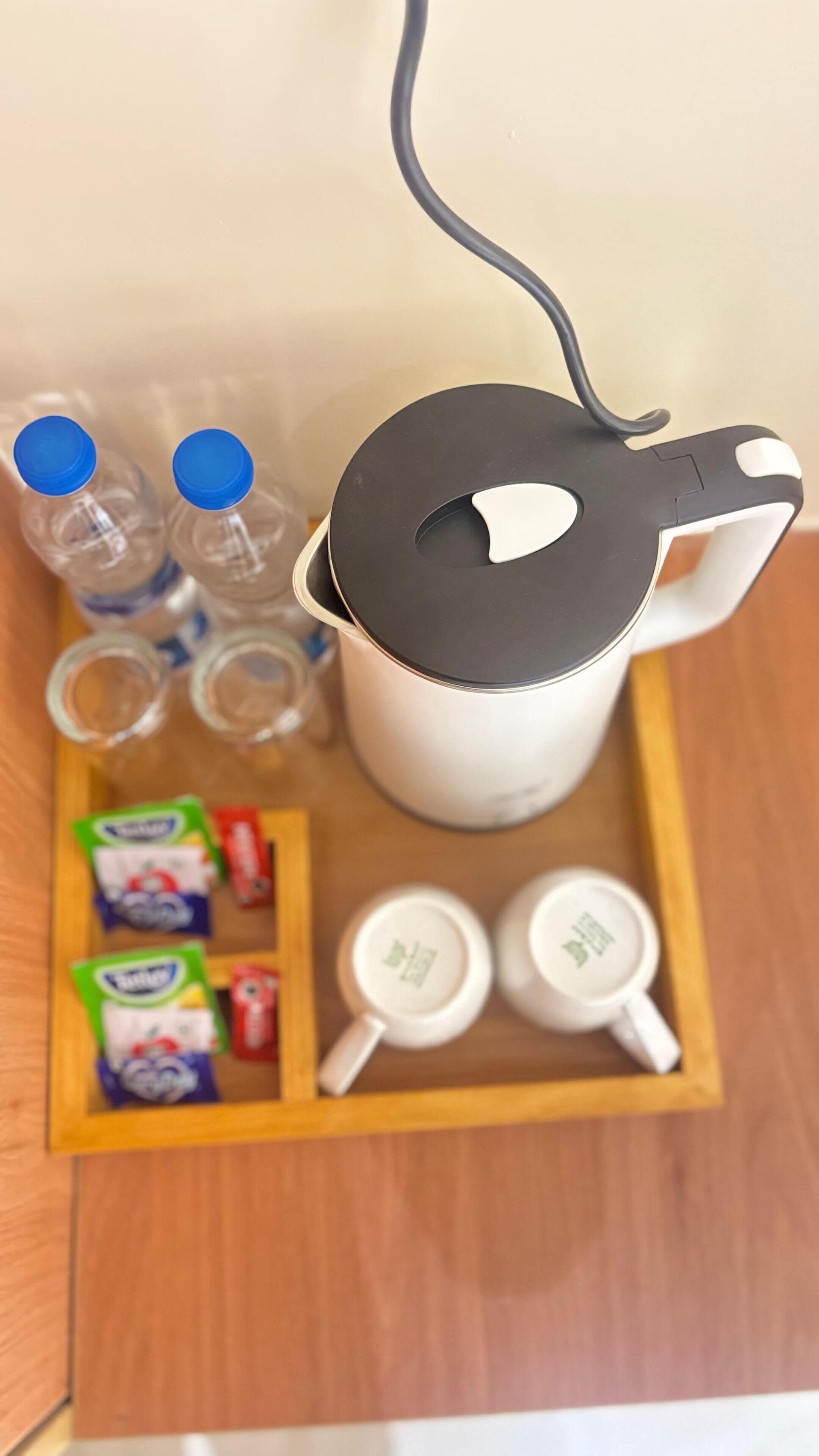 A white electric kettle with a black lid, two clear plastic bottles with blue caps, and a wooden tray with packets of tea and two white teacups.